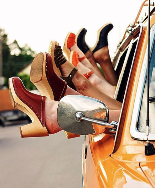 Four pairs of womens feet wearing high block heels sticking out the window of an orange car.